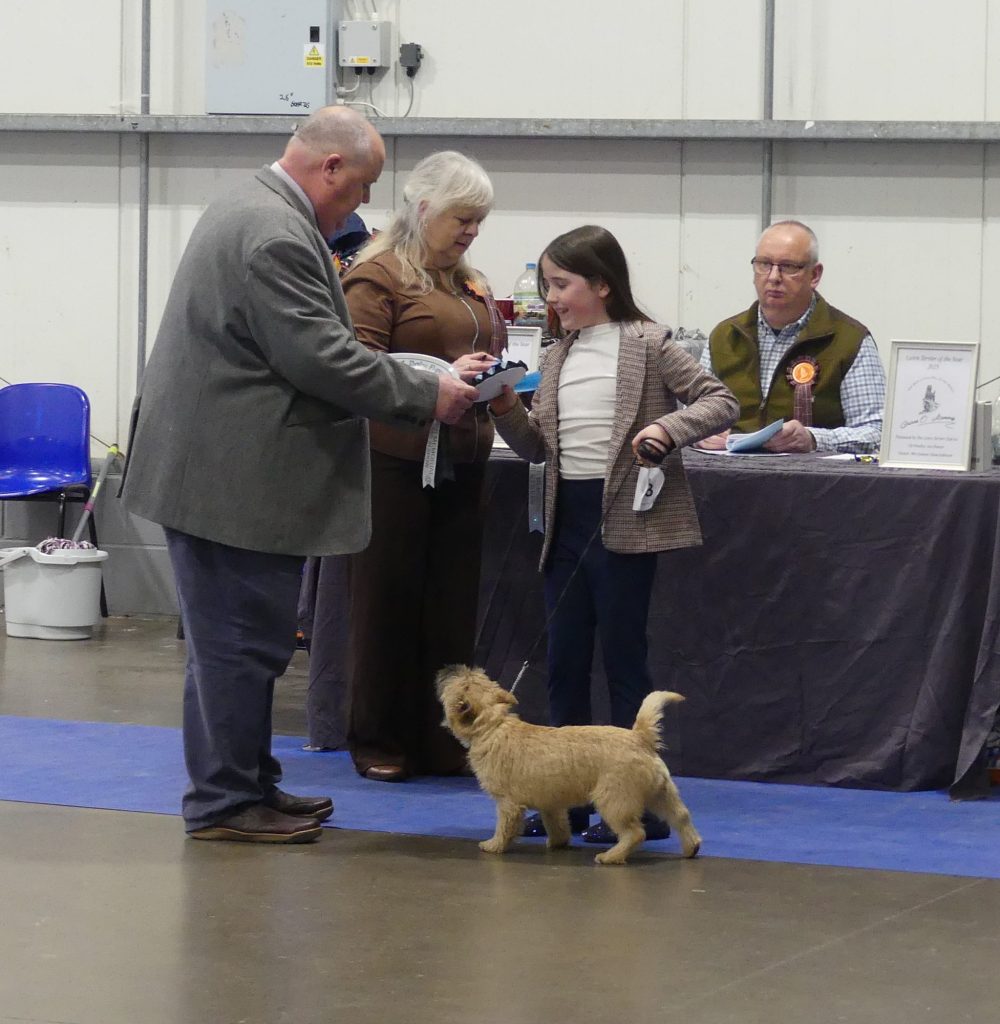 A young girl receiving a prize at a dog show, accompanied by a small dog, with judges and spectators in the background.