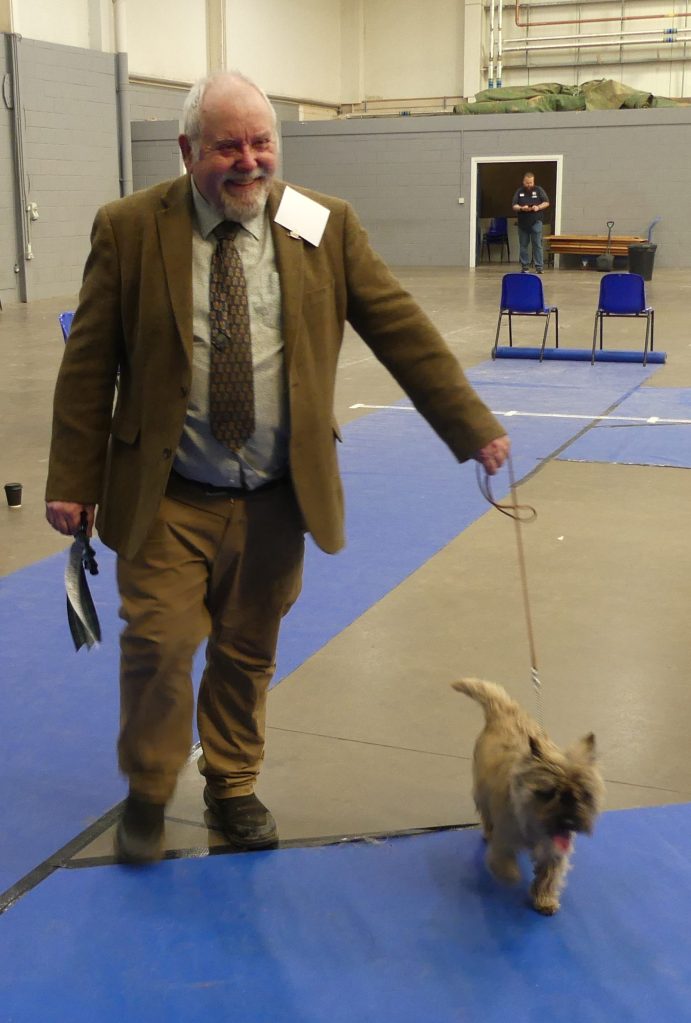 A smiling older gentleman in a brown blazer and tie walking with a small dog on a leash in a competition setting.